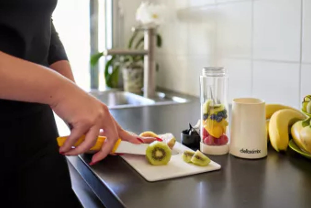 "Detoximix mini gebroken witte blender met twee transportflessen en receptenboek, ideaal voor gezonde smoothies onderweg. Dame snijdt vers fruit aan voor de blender."